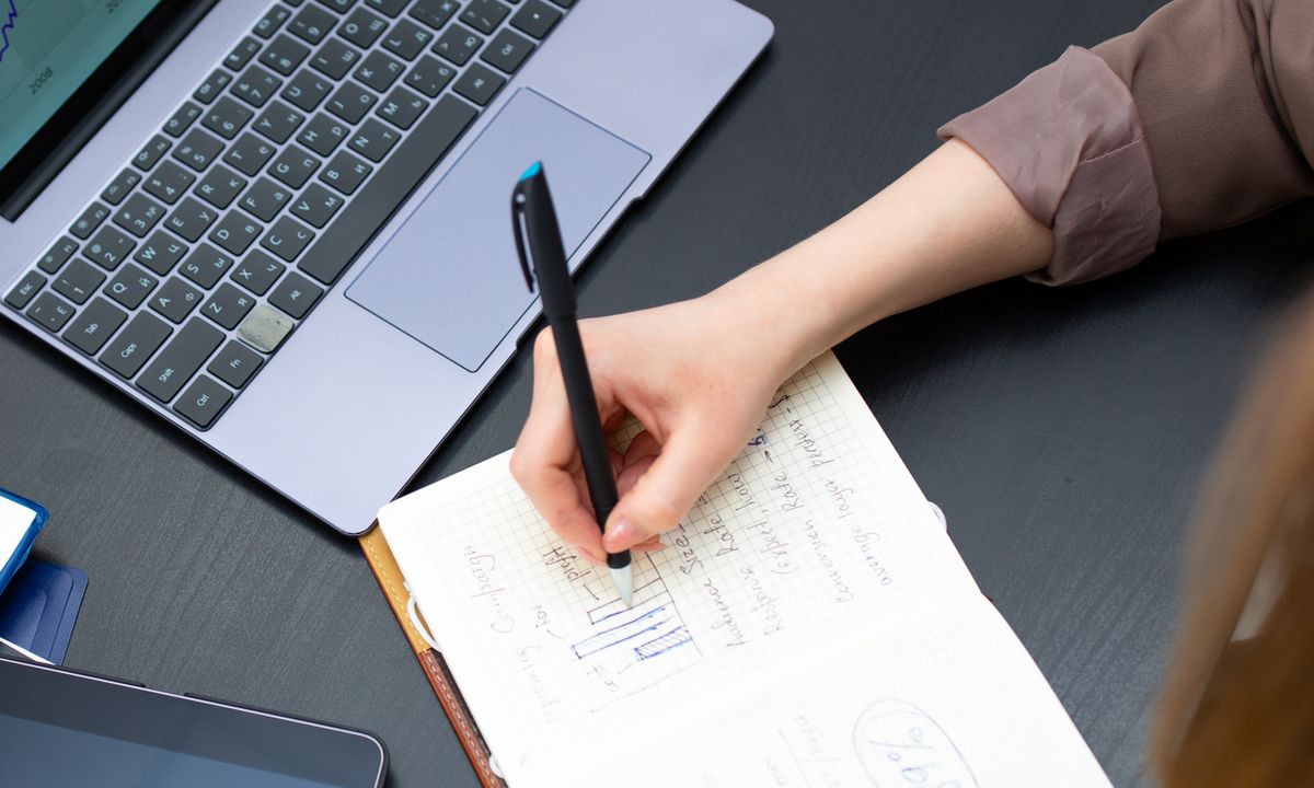 Hands writing notes beside a laptop and a cup of coffee